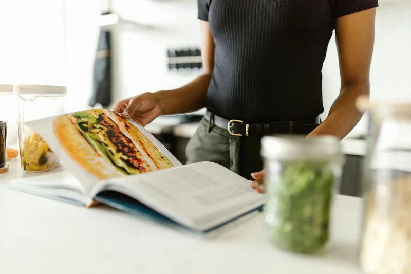 A person in a bright kitchen holds open a cookbook showing a large sandwich with meat, greens, and sauce on a white counter beside pasta containers.