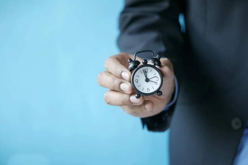 A person in a dark suit holds a miniature twin-bell alarm clock with a white dial and black numerals against a light blue background, symbolizing meal timing and eating schedule.