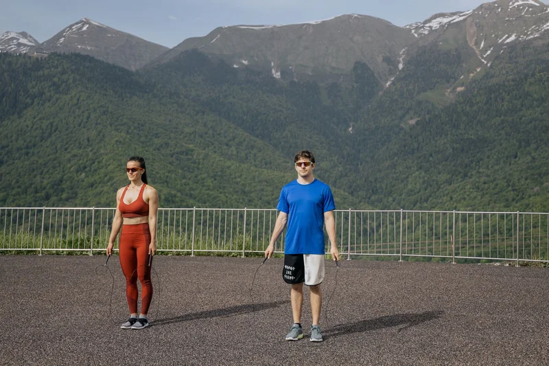 Two adults skipping rope outdoors with mountains in the background, showcasing fitness and nature.