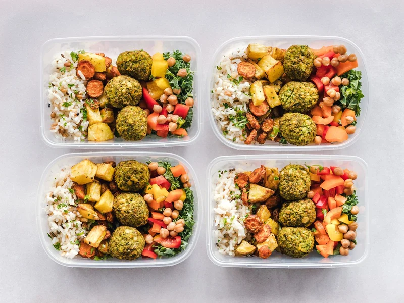 Four clear meal-prep containers hold portioned lunches with rice, roasted vegetables, falafel, chickpeas, and kale arranged on a light counter.